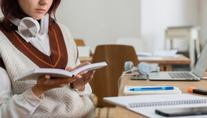 Zu sehen ist eine junge Frau, die eine Facharbeit geöffnet in der Hand hält und darin liest. Sie sitzt an einem Schreibtisch