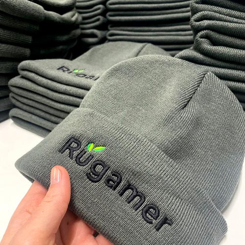 zu sehen ist eine grau bestickte Mütze im Vordergrund, im Hindergrund weitere Mützen, bestickt mit dem Logo der Firma Rügamer