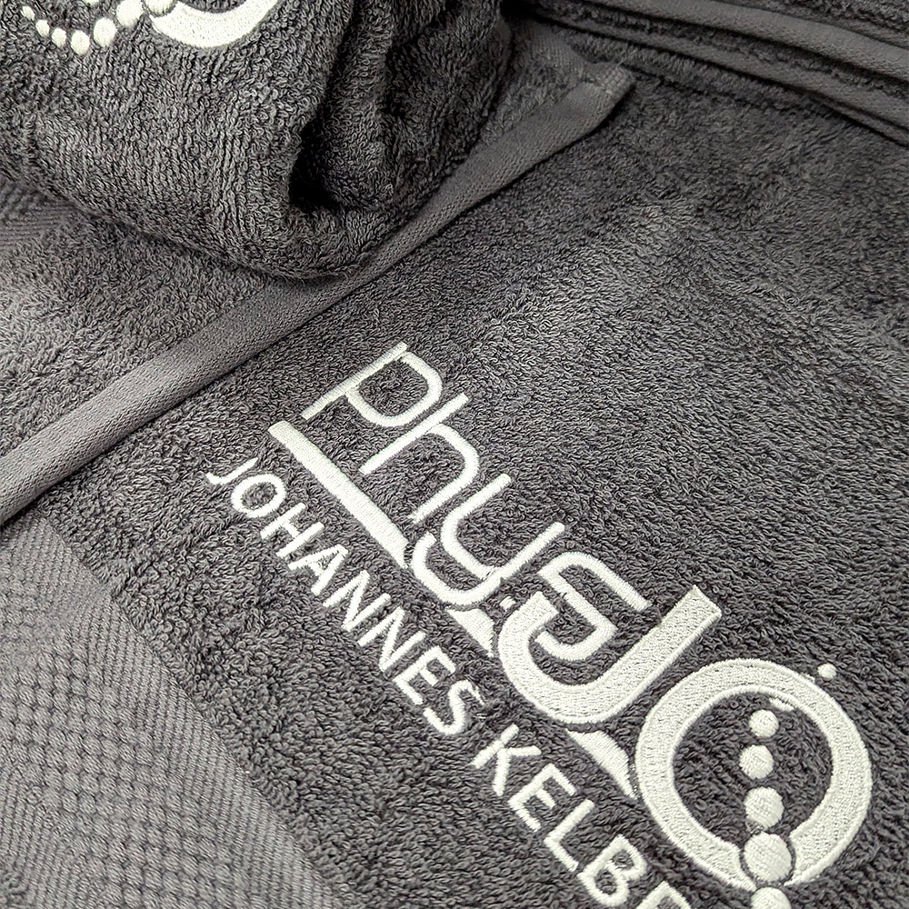 Zu sehen ist ein graues besticktes Handtuch mit einer weißen Aufschrift