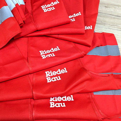 Zu sehen sind Warnwesten bedruckt in rot mit der weißen Aufschrift "Riedel Bau"