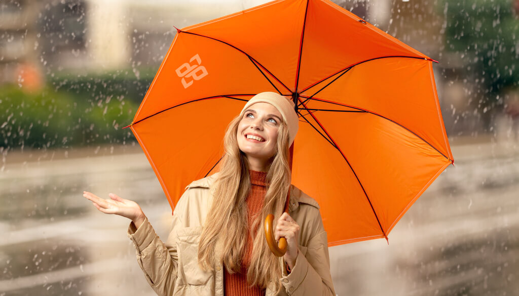 Das Bild zeigt eine blonde Frau mit einen unserer bedruckte Regenschirme in der Hand. Er ist geöffnet und hat die Farbe Orange. Die Frau hält ihre rechte Hand prüfend in den Regen.