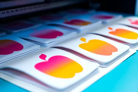 Das Bild zeigt bunte Karten, die sich in einem UV Druck Prozess befinden. Es werden orange-rote Äpfel gedruckt