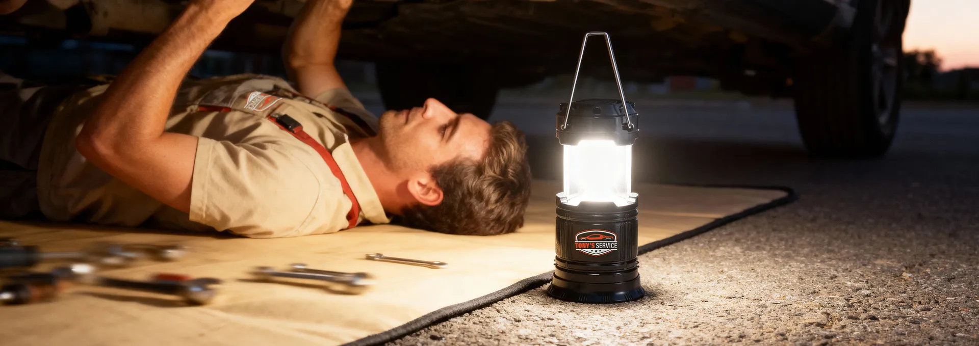 Mechaniker arbeitet im Dunkeln unter einem Fahrzeug, beleuchtet durch eine helle Arbeitslampe mit Logo ""Tony's Service"