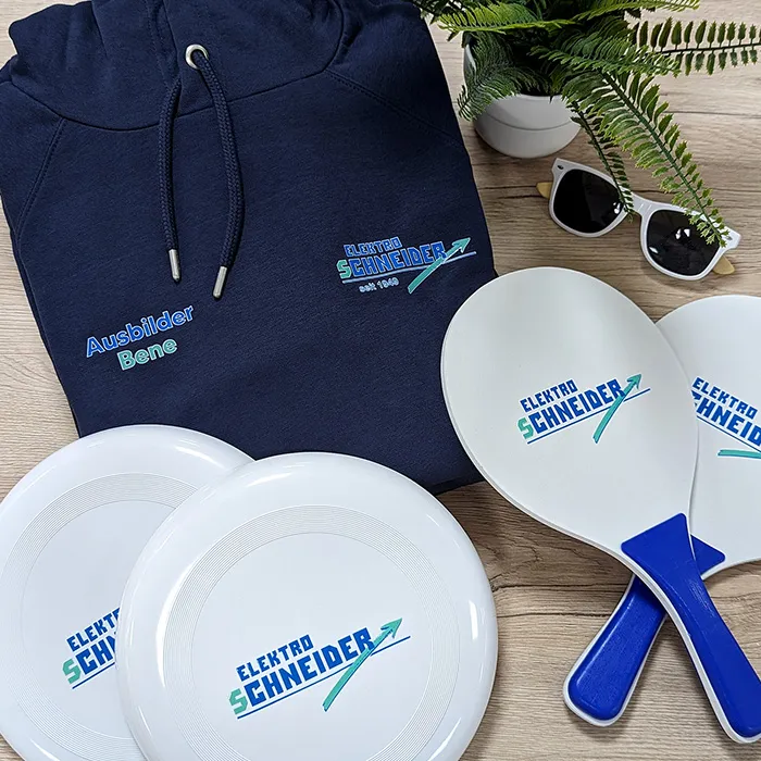 Sammlung von Werbeartikeln von Elektro Schneider in Schweinfurt, darunter Frisbees, Hoodie, Brille und Strandtennis-Schläger, auf Holzuntergrund arrangiert.