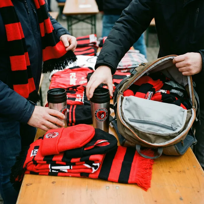 Hände ordnen Fanartikel mit Vereinslogo, darunter ein geöffneter Rucksack.