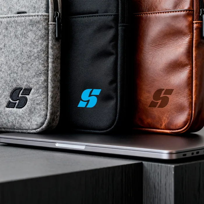 Nahaufnahme von drei Laptoptaschen in Grau (Filz), Schwarz (Textil) und Braun (Leder), jeweils mit dem Firmenlogo versehen.