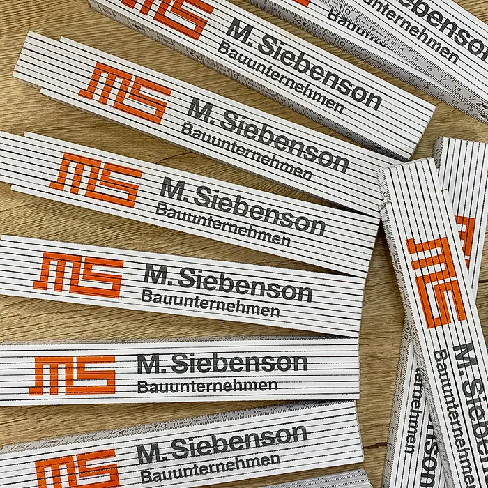 Personalisierte Arbeitskleidung für Siebenson – Bauunternehmen aus Schweinfurt 3 Weisse Zollstöcke mit dem Logo "Siebenson".