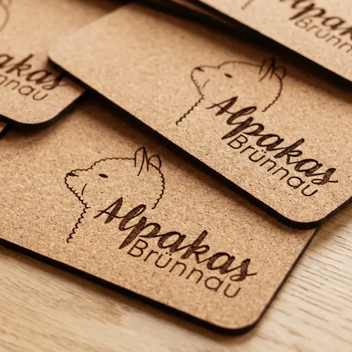Nahaufnahme von gestapelten, gelaserten Kork-Mousepads mit dem Logo "Alpakas Brünnau" und einer stilisierten Alpakazeichnung.