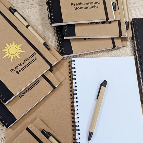 zu sehen ist Notizbuch bedrucken als Ringbuch in hellbraun und schwarz mit Stift, einer gelben Sonne und einem schwarzen Schriftzug