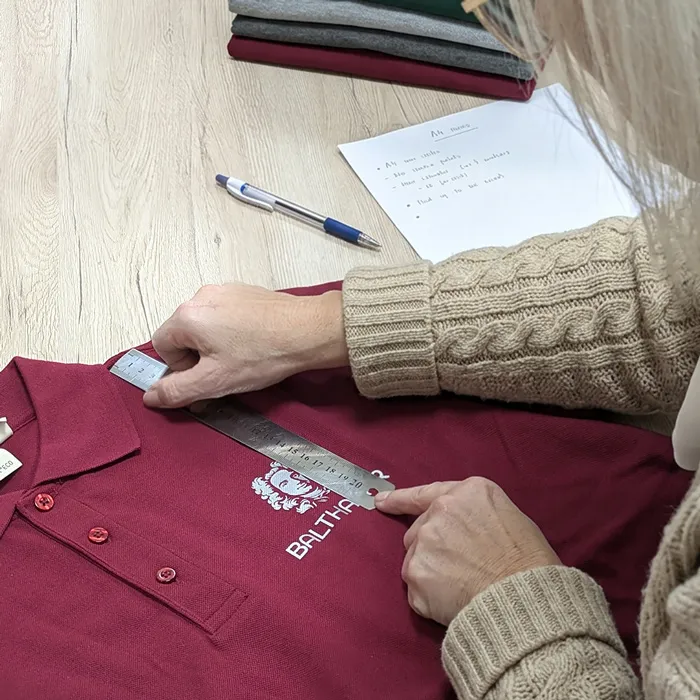 Mitarbeiterin misst mit einem Lineal die Position des weißen BALTHASAR-Logos auf einem bordeauxroten Poloshirt, vorbereitet für die Textilveredelung von Lamasuli.