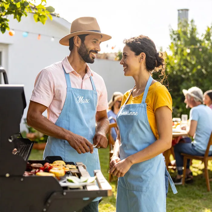 zu sehen sin Menschen mit bedruckte Grillschürze in hellblau und weißem Logo beim Grillen im Garten