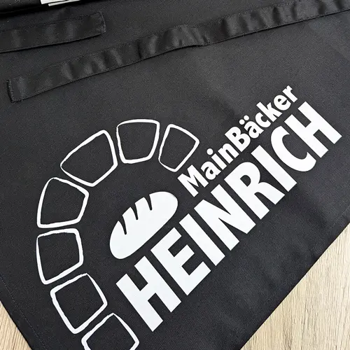 zu sehen ist eine bedruckte Schürze in schwarz mit der weißen Aufschrift "Main Bäcker Heinrich"