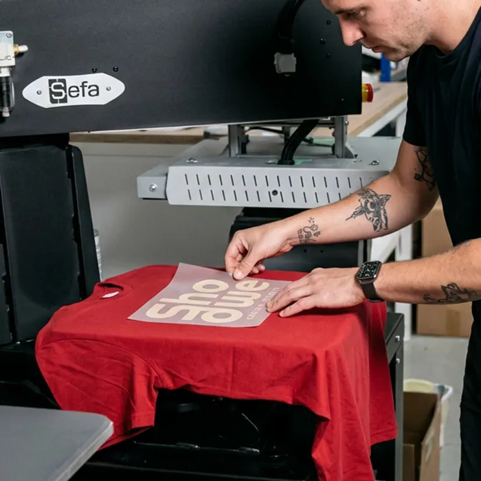 Mitarbeiter platziert eine Transferfolie mit weißem Schriftzug auf einem roten T-Shirt unter einer Transferpresse, vorbereitet für den Textildruck von Lamasuli.