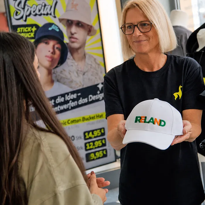Mitarbeiterin präsentiert eine weiße Cap mit grün-orangem RELAND-Logo im Textildruckgeschäft, individuell bedruckt von Lamasuli.